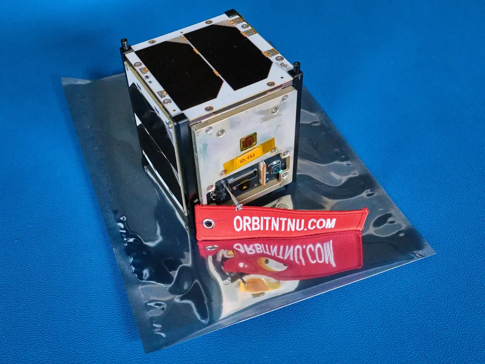 FramSat-1, en liten kubeformet satellitt med solcellepaneler, på en antistatisk pose med røde Orbit NTNU-merkelapper